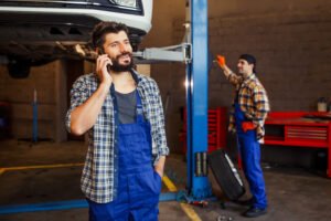 smiling mechanic talking smartphone with lifted car coworker | Roberts Mobile Mechanics